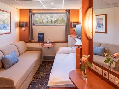 Geräumige Kabine des Flusskreuzfahrtschiffs Bijou du Rhône mit Sofa, Einzelbett und Fenster. Ein Spiegel und ein kleiner Tisch mit Getränken ergänzen die Ausstattung.