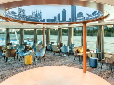 Lounge des Flusskreuzfahrtschiffs Bijou du Rhône. Viele Sessel und Tische. Fenster zeigen Fluss. Deckenpaneel mit Stadtpanorama-Mural. Rundes Podest im Vordergrund.