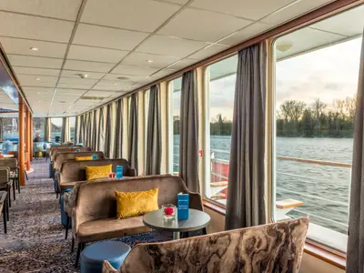 Blick in die helle Lounge der Bijou du Rhône. Reihen bequemer Sofas säumen große Fenster mit Aussicht auf den Fluss und bewaldetes Ufer. Tische sind mit Barkarten bestückt.