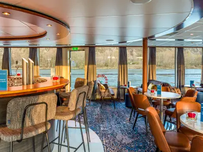 Innenansicht der Lounge-Bar auf der Bijou du Rhône. Sie zeigt Sitzbereiche, eine Bar und Panoramafenster mit Blick auf den Fluss und grüne Ufer. Helles Sonnenlicht durchflutet den Raum.
