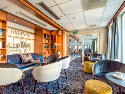Lounge-Bereich auf der Bijou du Rhône. Man sieht Sessel, Sofa, Bücherregale, 'LIBRAIRIE'-Schriftzug und große Fenster. Im Hintergrund ist ein Barbereich erkennbar.
