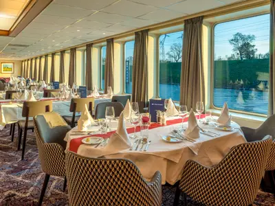 Der Speisesaal der Bijou du Rhône mit elegant gedeckten Tischen. Große Fenster bieten Blick auf den Fluss und Uferlandschaft, durchflutet von Abendlicht.