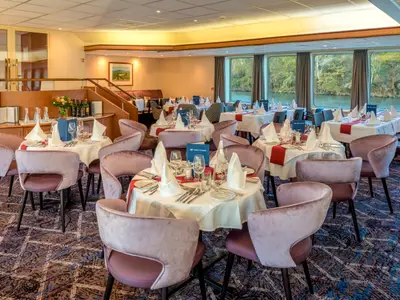 Innenansicht des Restaurants auf dem Flusskreuzfahrtschiff Bijou du Rhône mit gedeckten Tischen und Samtstühlen. Große Fenster bieten einen Blick auf den Fluss und bewaldete Ufer. Der Raum ist hell beleuchtet und elegant eingerichtet.