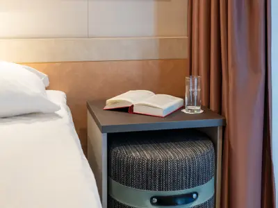 Nahaufnahme einer modern eingerichteten Kreuzfahrtkabine. Ein frisch gemachtes Bett, ein Nachttisch mit einem aufgeschlagenen Buch und einem leeren Trinkglas stehen bereit.
