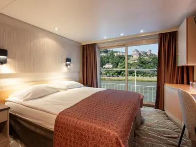 Helle Kabine der Bolero mit bequemen Betten und Schreibtisch. Die Fensterfront bietet Panoramablick auf einen Fluss und eine alte Burg auf einem Hügel.