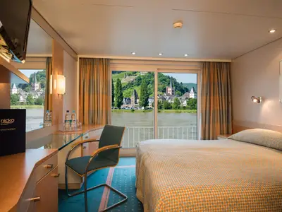 Helle Kabine der nicko cruises Bolero mit Bett, Schreibtisch und Panoramafenster. Der Ausblick zeigt einen Fluss, ein charmantes Dorf mit Kirchturm und grüne Weinhänge.
