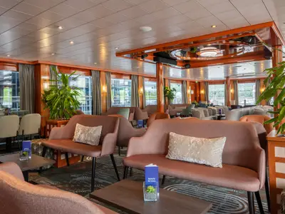 Lounge des Flusskreuzfahrtschiffs Bolero. Komfortable Sitzgruppen, große Fenster, helle Decke mit Holzakzenten und Pflanzen. Einladendes Interieur für Gäste.