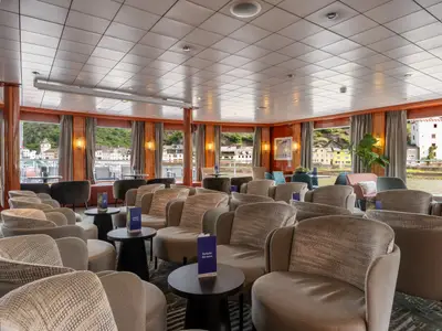 Innenansicht der Panoramalounge des Flusskreuzfahrtschiffs Bolero mit vielen Sesseln und Tischen. Durch die Fenster blickt man auf eine Uferlandschaft mit Häusern und Hügeln am Fluss.