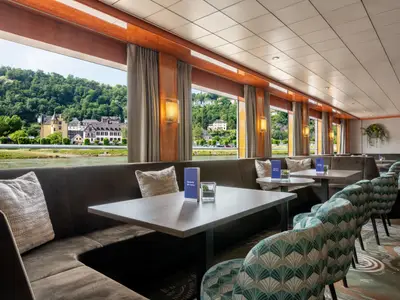 Innenansicht des Panoramarestaurants der Bolero mit Blick auf Fluss, Hügel und Gebäude. Dunkle Sitzbänke, Tische und gemusterte Stühle sind zu sehen.
