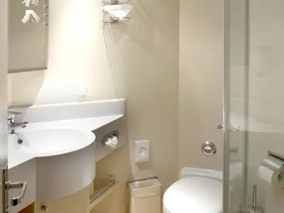 Detailansicht eines modernen, hellen Badezimmers einer Kreuzfahrtkabine. Es zeigt ein weißes Waschbecken mit Chromarmatur, eine Toilette und eine Duschkabine mit Glaswand. Beige Wände und weiße Sanitärobjekte prägen das Bild.
