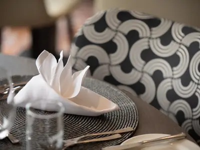Nahaufnahme eines gedeckten Tisches in einem Restaurant an Bord. Eine weiße, kunstvoll gefaltete Stoffserviette liegt auf einem runden, dunkelgrauen Platzset, umgeben von Besteck und einem Trinkglas. Im unscharfen Hintergrund ist ein Stuhl mit schwarzem und weißem geometrischem Bogenmuster zu sehen.