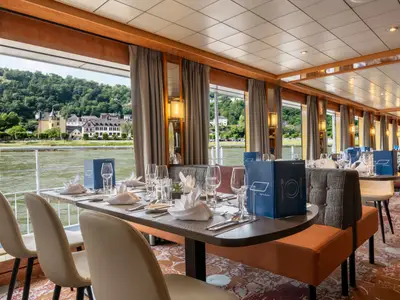 Das helle Restaurant des Flusskreuzfahrtschiffs Bolero mit gedeckten Tischen. Große Panoramafenster bieten eine Aussicht auf grüne Ufer und historische Häuser am Fluss.