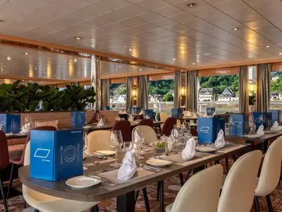 Das helle Bordrestaurant des Flusskreuzfahrtschiffs Bolero ist mit gedeckten Tischen und Stühlen ausgestattet. Durch die Fenster ist eine grüne Flusslandschaft mit Uferbebauung zu sehen. Blaue Menümappen liegen aus.