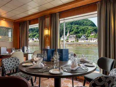Restaurant auf dem Flusskreuzfahrtschiff Bolero. Gedeckte Tische mit Blick auf einen Fluss und ein Dorf am Ufer mit Kirche und bewaldeten Hügeln.