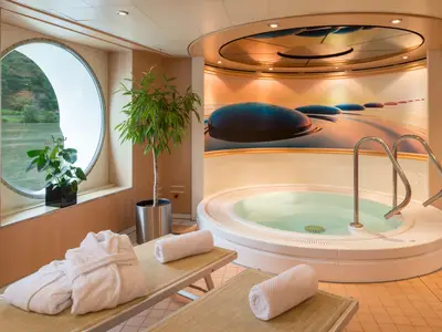 Der Wellnessbereich der Bolero zeigt einen runden Whirlpool und zwei Liegen mit Bademänteln. Ein großes Bullauge bietet Blick auf die vorbeiziehende Flusslandschaft.