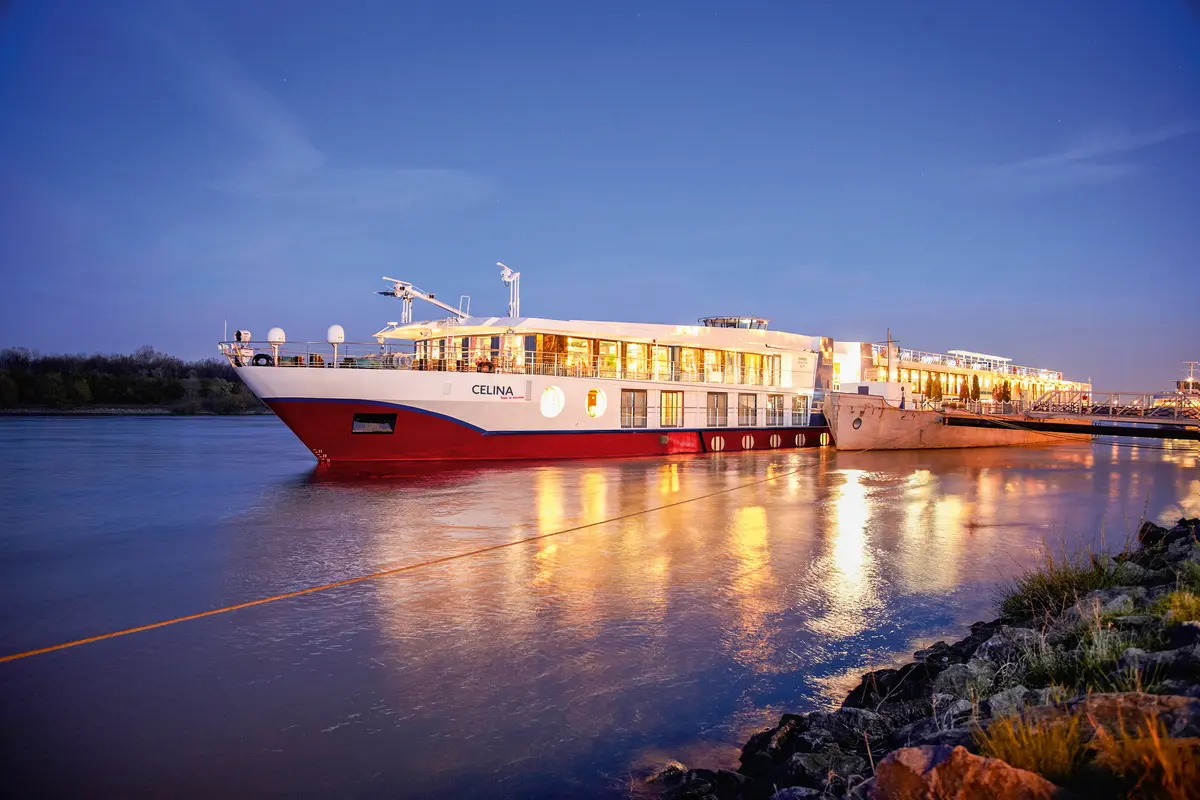 Das Flussschiff Celina liegt beleuchtet am Flussufer. Die roten und weißen Rumpffarben spiegeln sich im ruhigen Wasser unter einem blauen Abendhimmel wider.
