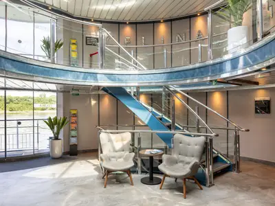 Die helle Lobby der Celina mit blauer Treppe, Glasgeländern und zwei grauen Sesseln. Große Fenster bieten Blick auf den Fluss und die grüne Uferlandschaft.