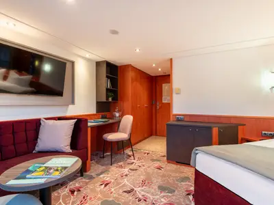 Die Kabine der Celina zeigt ein rotes Sofa mit TV, Schreibtisch und Bett. Der Raum hat floralen Teppich und Holzelemente.