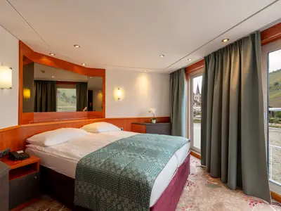 Komfortable Kabine der Celina mit Doppelbett, Holzeinrichtung und großem Fenster. Blick auf eine Flusslandschaft mit grünen Hügeln und einer Kirche.