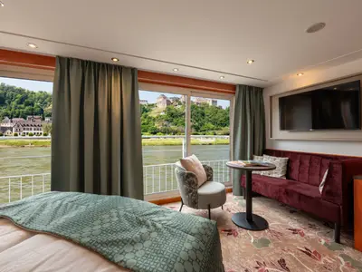 Eine helle Kabine der nicko cruises Celina mit bodentiefen Fenstern bietet Ausblick auf eine Flusslandschaft mit Burg und Häusern. Innen sind ein Bett, ein Sessel, ein rotes Sofa und ein TV zu sehen.