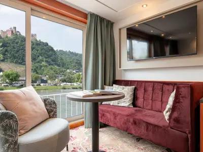 Innenansicht einer modernen Kabine der Celina mit rotem Samtsofa, Sessel und Beistelltisch. Durch die großen Fenster blickt man auf eine grüne Flusslandschaft mit einer historischen Burg auf einem Hügel.