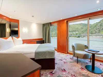 Helle Kabine an Bord der Celina mit Bett, Sessel, Tisch und großer Fensterfront. Man blickt auf einen Fluss und bewaldete Hügel. Ein floraler Teppich und orangefarbene Holzpaneele prägen das Interieur.