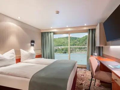 Eine Kabine der nicko cruises Celina mit Bett, Schreibtisch und TV. Der Blick aus dem Fenster zeigt eine grüne Flusslandschaft, Hügel und Uferbebauung. Helle, moderne Einrichtung.