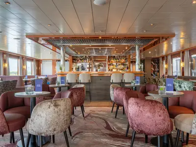 Blick in den Bar- und Loungebereich der Celina. Bequeme Sitzgelegenheiten, eine gut gefüllte Bar mit Hockern und große Fenster zur Außenseite.