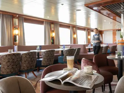 Eine gemütliche Lounge an Bord der Celina zeigt Sitzbereiche, Tische mit Kaffee und Magazinen. Ein Kellner bewegt sich im Hintergrund, während Tageslicht durch die Fenster fällt.
