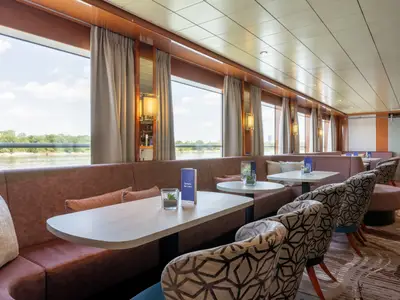 Der helle Loungebereich an Bord der Celina zeigt Fenster mit weitem Flussblick, gepolsterte Bänke, Tische und Sessel mit gemustertem Bezug.