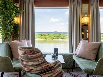 Ein heller Sitzbereich an Bord der Celina mit bequemen Sesseln und einem Beistelltisch. Durch die großen Fenster blickt man auf einen Fluss, bewaldete Ufer und einen blauen Himmel mit Wolken.