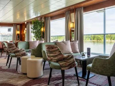 Heller Salon auf der Celina mit Sitzgruppen aus grünen Sesseln und Sofas. Große Fenster bieten Ausblick auf einen Fluss und bewaldetes Ufer. Moderne, einladende Ausstattung.