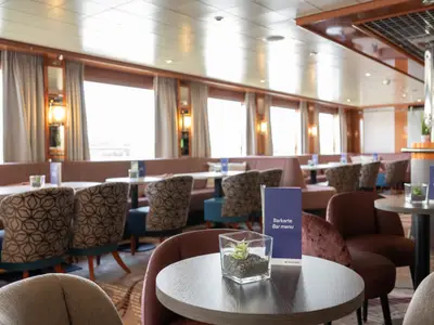 Die elegante Lounge des Flusskreuzfahrtschiffs Celina präsentiert gemütliche Sitzgruppen entlang großer Fenster, ergänzt durch eine stilvolle Bar. Menüs und Dekorationen sind auf den Tischen sichtbar.