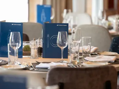 Ein gedeckter Tisch im Restaurant der nicko cruises Celina zeigt blaue Menükarten, Gläser, Besteck, Teller und Salz-Pfefferstreuer. Im unscharfen Hintergrund sind weitere Tische erkennbar.