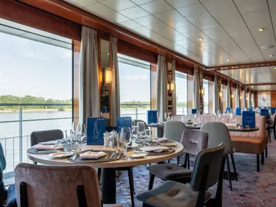 Innenansicht des Restaurants auf der Celina. Gedeckte Tische mit Besteck und blauen Menükarten. Große Fensterfront mit Ausblick auf den Fluss und die Uferlandschaft.