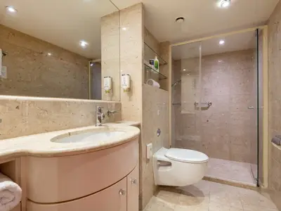 Modernes Badezimmer an Bord der Celina. Es zeigt Waschtisch, Spiegel, wandhängendes WC und eine Duschkabine. Helle Steinfliesen an Wänden und Boden.