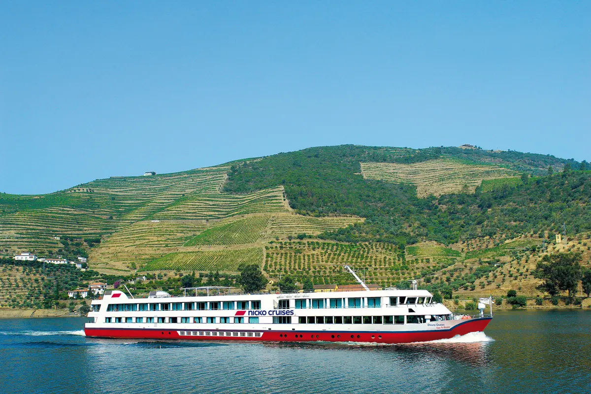 Weiß-rotes Flussschiff mit dem Schriftzug "Nicko Cruises" und "Douro Queen" fährt auf einem blauen Fluss. Im Hintergrund erheben sich grüne, terrassierte Weinberge unter einem klaren blauen Himmel.