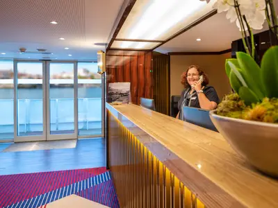 Die Rezeption der Douro Queen mit lächelnder Mitarbeiterin, die telefoniert. Ein beleuchteter Tresen, große Fenster mit Blick auf den Fluss und eine weiße Orchidee prägen den Raum.