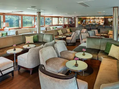 Die elegante Lounge der Douro Queen bietet komfortable Sofas und Sessel. Große Fenster zeigen den Fluss. Hinten rechts befindet sich eine Bar, an der ein Barkeeper arbeitet.