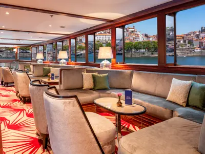 Blick in die gemütliche Lounge der Douro Queen: Helle Sofas und Sessel stehen entlang großer Fenster, die eine malerische Flusslandschaft mit bunten Häusern am Ufer zeigen.