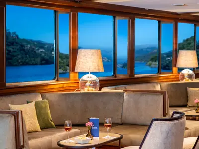 Abendlicher Salon der Douro Queen mit Sofas, Tischen, Wein. Blick durch die Fenster auf eine ruhige Flusslandschaft in der Dämmerung.