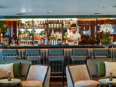 Bar der Douro Queen. Ein Barmann mixt einen Cocktail. Hinter der Theke Flaschenregale, davor gemütliche Loungemöbel. Elegante Einrichtung mit Orchideen.