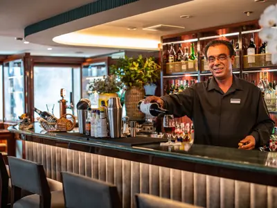 Ein lächelnder Barkeeper der Douro Queen schenkt an der Bar Rotwein ein. Im Hintergrund sind Spirituosenregale und auf der Theke Barutensilien sichtbar.