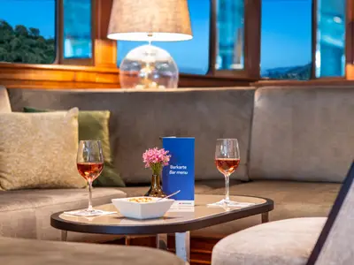 Gemütlicher Barbereich der Douro Queen bei Abendstimmung. Zwei Gläser Roséwein, Snacks, Barkarte auf Tisch. Sofas und Blick auf dunkle Hügellandschaft.