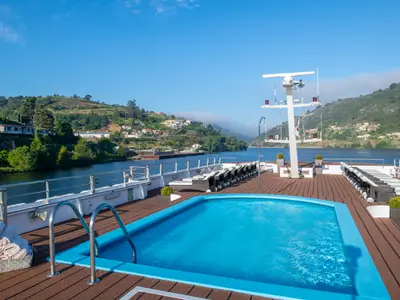 Blick vom Oberdeck der Douro Queen auf einen Pool und Liegestühle. Der Fluss und grüne, bebaute Hügel sind unter blauem Himmel sichtbar.