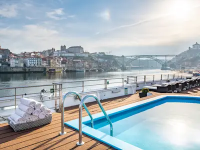 Das Pooldeck der Douro Queen mit blauem Pool und Handtüchern. Dahinter der Fluss Douro mit Porto und markanter Bogenbrücke.