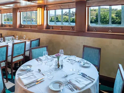 Das Restaurant der Douro Queen bietet gedeckte Tische mit blauen Stühlen. Durch die Fenster blickt man auf die Douro-Flusslandschaft mit grünen Hängen und Weinbergen.
