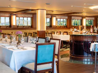 Innenansicht des Restaurants der Douro Queen. Gedeckte Tische mit Gläsern und Blumen. Fenster mit Blick auf grüne Hügel. Eine Bar mit Weinen und Sektkühler ist rechts zu sehen.