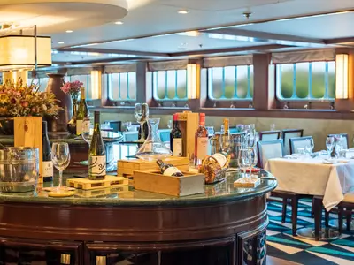 Gastronomiebereich der Douro Queen. Eine Theke präsentiert Weine und Gläser. Im Hintergrund sind stilvoll gedeckte Tische und Bullaugenfenster sichtbar.