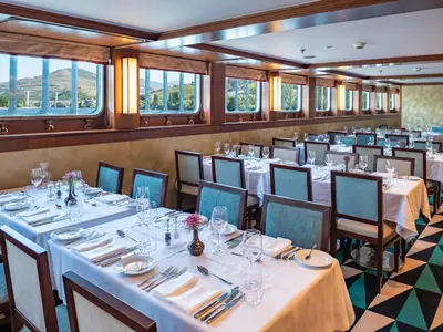 Ein elegantes Restaurant mit vielen weiß gedeckten Tischen und türkisfarbenen Stühlen. Große Fenster bieten einen malerischen Blick auf grüne Weinberge und einen Fluss mit Brücken.